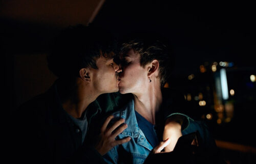 Young gay couple in casual clothing kissing while holding mobile phone on balcony at night